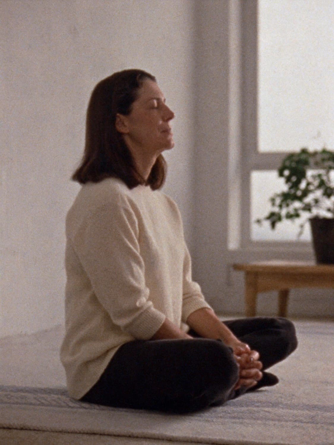 A woman sitting cross-legged by a window with her eyes closed